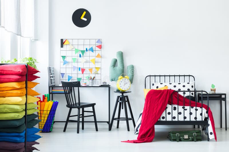 Pillows in bright child`s room. Colorful pillows in bright child`s room with red blanket on the bed and yellow clock on a stool near desk with chair royalty free stock photos