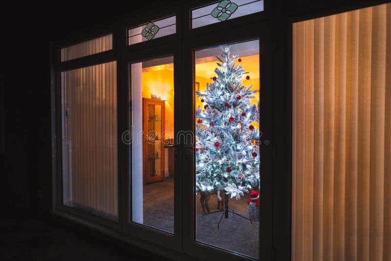 A frosted white artificial christmas tree with red and silver decorations seen through large patio doors with vertical slat blinds. And bevel window designs stock photos