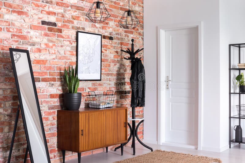 Mirror next to wooden cabinet in entrance hall interior with white door and poster on red brick wall. Real photo stock photos