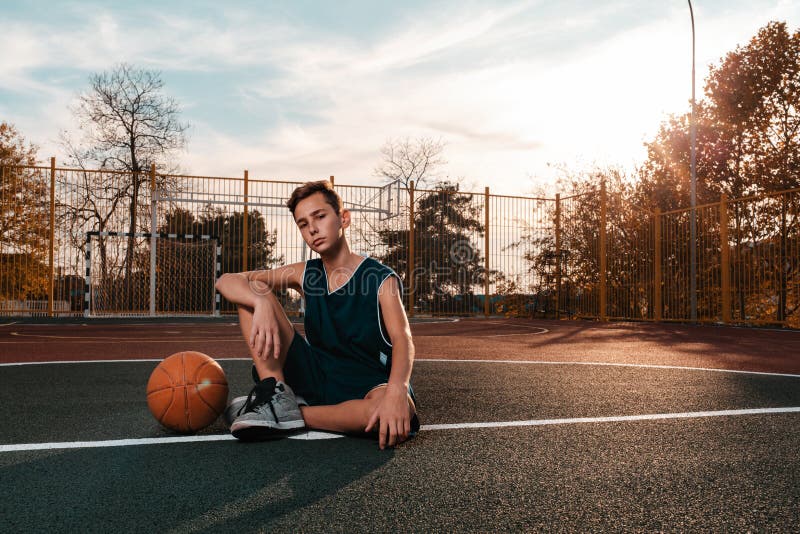Sports and basketball. A young teenager in a black tracksuit poses with a basketball while sitting on the ground. Sunset. Copy royalty free stock image