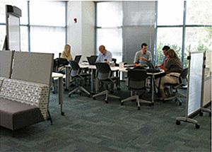 A long table positioned along a wall of windows and separated from workstations where workers are meeting in a small group at one end and individuals are working on their laptops on the other end