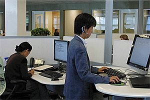 A woman seated in front of a computer and man standing at a workstation on a computer while wearing a Bluetooth headset